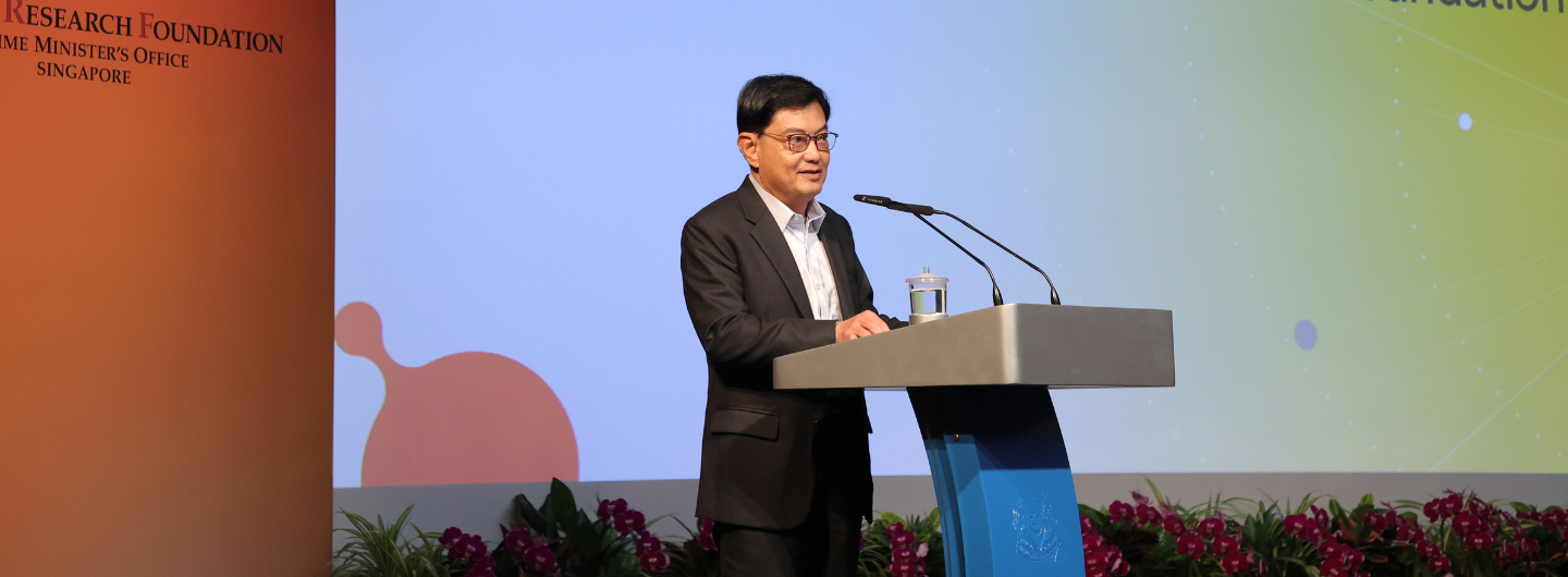 Man at podium with microphones, water glass, Research Foundation Singapore logo.